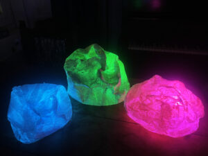 Three paper mache stones, lit up in bright colours, in a dark room.