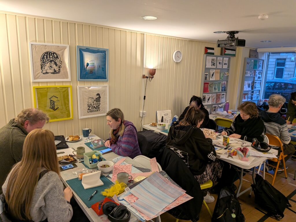 A busy workshop scene in a bright, cosy room where a group of people sit around tables making artist books and zines. The tables are covered with cutting mats, paper, pens, collage materials, cups, bowls of snacks, and works in progress. On the walls are framed zine-related prints, and shelves at the back hold pamphlets and booklets. The atmosphere feels focused and collaborative, with everyone engaged in making and conversation.