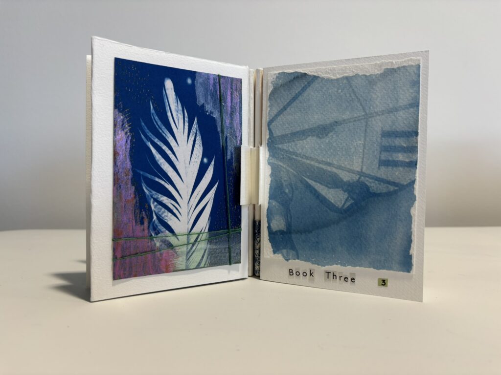 Small handmade artist’s book displayed open on a white surface against a plain grey background. The left page features a vivid blue and purple image with a white feather- or leaf-like form rising vertically, crossed by fine green thread. The right page shows a translucent blue-toned image on textured torn-edge paper, resembling an abstract photograph or shadowy structure. Along the bottom, cut-out text reads “Book Three” with a small number 3. The book has a delicate, tactile handmade quality.