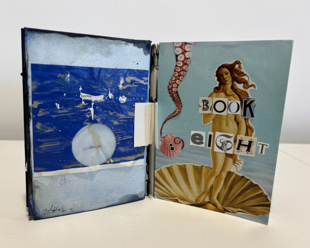 A handmade artist’s book is shown partially open on a white surface, revealing both the front cover and the inside back page. On the right, the front cover features a collage based on Botticelli’s Birth of Venus: a nude female figure with flowing auburn hair stands on a large scallop shell against a pale blue background. The title “BOOK EIGHT” is arranged across her body in mismatched cut-out letters, giving a ransom-note collage effect. An octopus tentacle curls down from the top left of the cover, ending near a small pink shell marked with the number 8. On the left, the inside back page is painted in layered shades of blue, with darker brushstrokes and gold-toned streaks suggesting sea or sky. A circular pale shell or disc is attached near the lower centre, with faint handwritten text across it. Torn fragments of printed text are embedded in the painted surface near the middle, partially obscured by the paint. The book’s black stitched spine and handmade binding are visible between the pages, emphasising its crafted, sculptural quality. The overall composition evokes marine imagery and classical references, with a strong oceanic blue palette.