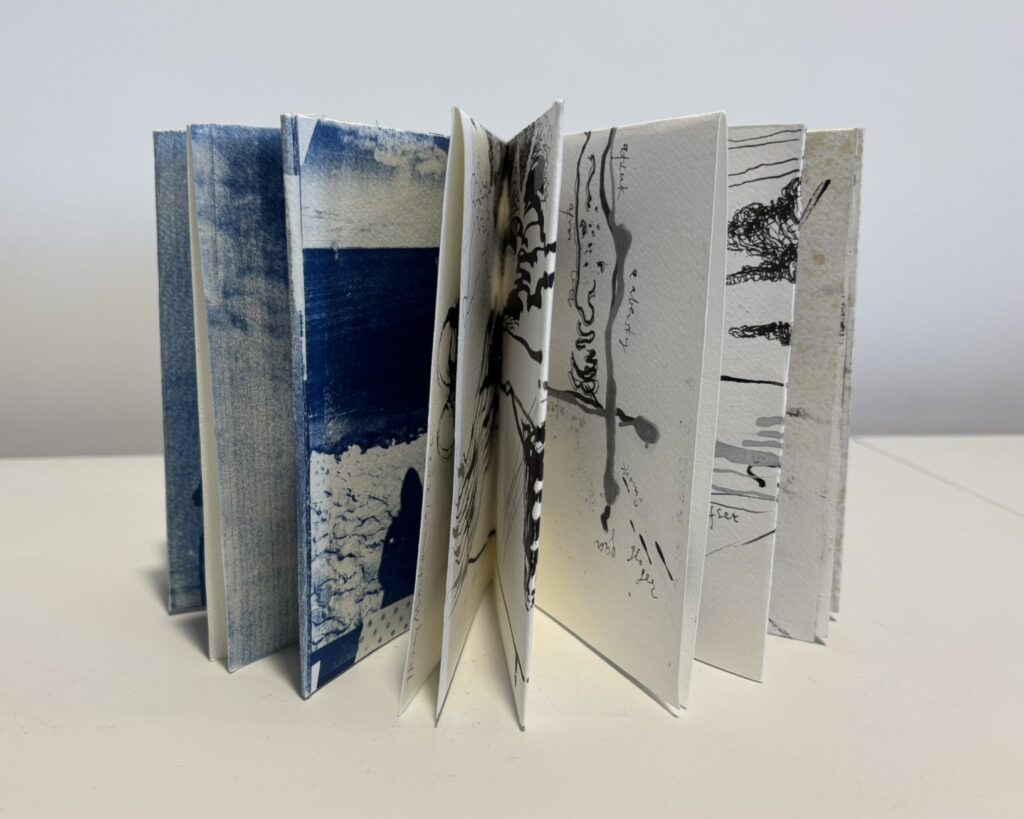 A handmade artist’s book stands upright with its pages fanned open in a sculptural display on a white surface. The pages spread in a zigzag, accordion-like formation, revealing multiple painted and collaged interiors. On the left, several pages are washed in layered blues ranging from pale sky blue to deep navy, evoking sea and sky. Toward the centre and right, black ink drawings, fluid brush marks, drips, and fragments of handwritten text appear across white textured pages. Some pages include abstract line work and organic shapes, while others feature blocks of darker blue collage or paint. The varied page surfaces and visible folds emphasise the tactile, handmade quality of the book, with each spread appearing as an individual artwork within the whole object.