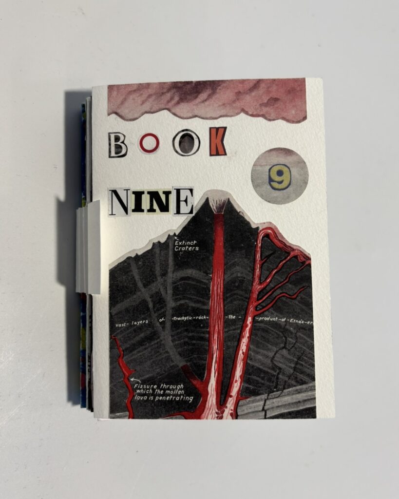 Front cover of a small handmade artist’s book titled “BOOK NINE,” with the words assembled from mismatched cut-out letters in a collage style. A circular cut-out on the right contains the number “9.” The main image is a vintage scientific illustration of a volcano cross-section, showing dark rock layers split by vivid red lava channels rising to the crater. Small printed labels identify features such as “extinct craters” and “fissure through which the molten lava is penetrating.” Along the top edge, a band of maroon watercolour suggests smoke or cloud. The book is photographed closed against a plain light background, with the thickness of the pages visible at the left edge.