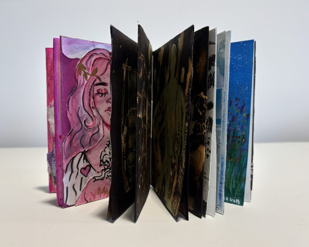 Small handmade artist’s book displayed standing with its pages fanned open in an accordion-like arc. The visible pages reveal a sequence of contrasting artworks: at the left, a pink-toned illustrated portrait of a person with long hair; in the centre, several dark black pages with gold and earthy markings; and toward the right, lighter white and bright blue pages with painted and printed imagery. The fan arrangement shows the rhythm and variety of the book’s interior, moving from vivid pinks through shadowy blacks to cooler blues and whites. The book is photographed against a plain light background, emphasising the sculptural quality of the open pages.