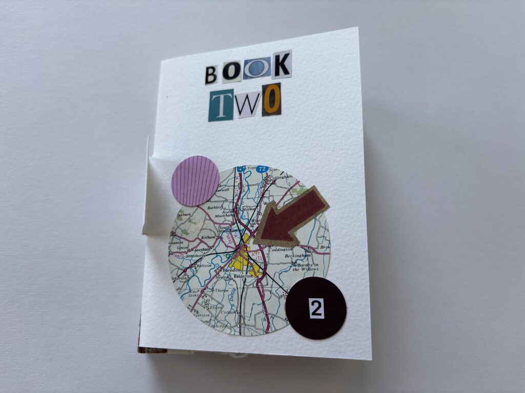 A hand-bound book. The cover is collaged, with letters spelling out "Book Two". There is a circle cut from a map, with an arrow pointing to a junction at the centre. There are two smaller circles, one pink, the other black. The black circle contains a numeral 2.