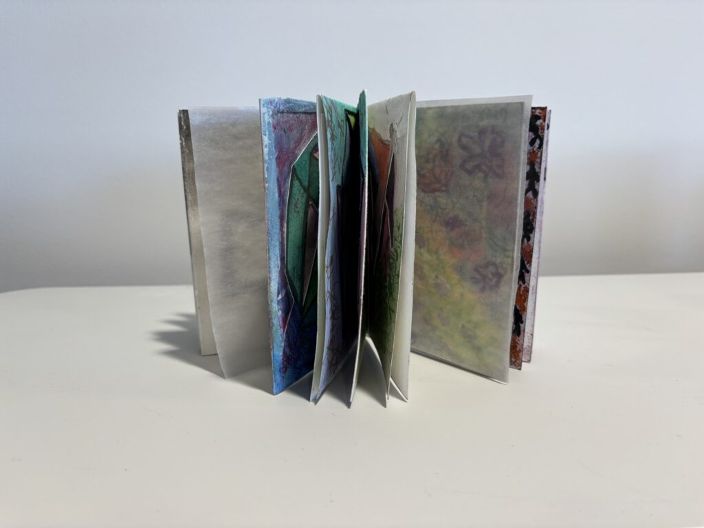 The book stands upright with pages fanned open, revealing multiple layers of mixed-media spreads in translucent and opaque papers. Glimpses of bright blues, greens, pinks, floral motifs, and geometric forms can be seen through the staggered pages, creating a sculptural, accordion-like silhouette. The overlapping translucent sheets allow colours and shapes from different pages to softly show through, highlighting the book’s tactile, handmade, and dimensional quality.