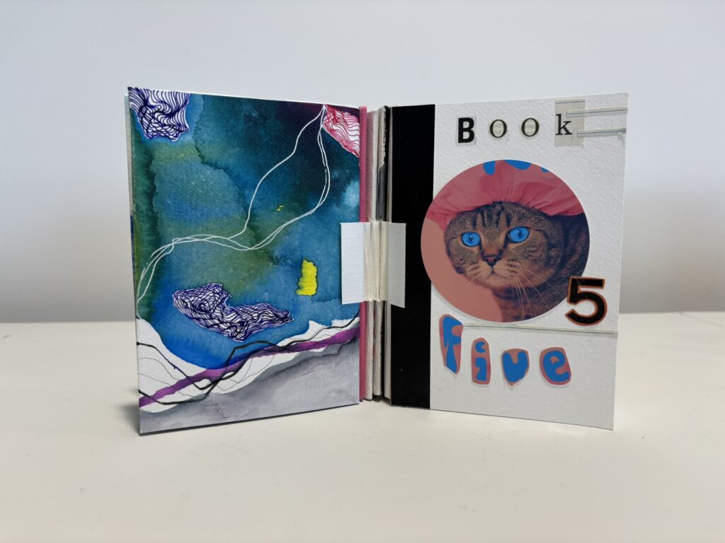 The book is shown partially open to its title spread. On the right page, cut-out letters spell “Book 5 Five” around a circular image of a cat with striking bright blue eyes wearing a pink ruffled bonnet. The page uses a bold collage style with black vertical bands and silver tape-like accents. The left page is an abstract mixed-media composition in rich blues, teals, purple, black, and white, with fluid ink-like washes, fine contour-style line drawings, and a small bright yellow shape. A folded white paper hinge at the centre highlights the handcrafted binding and interactive structure.