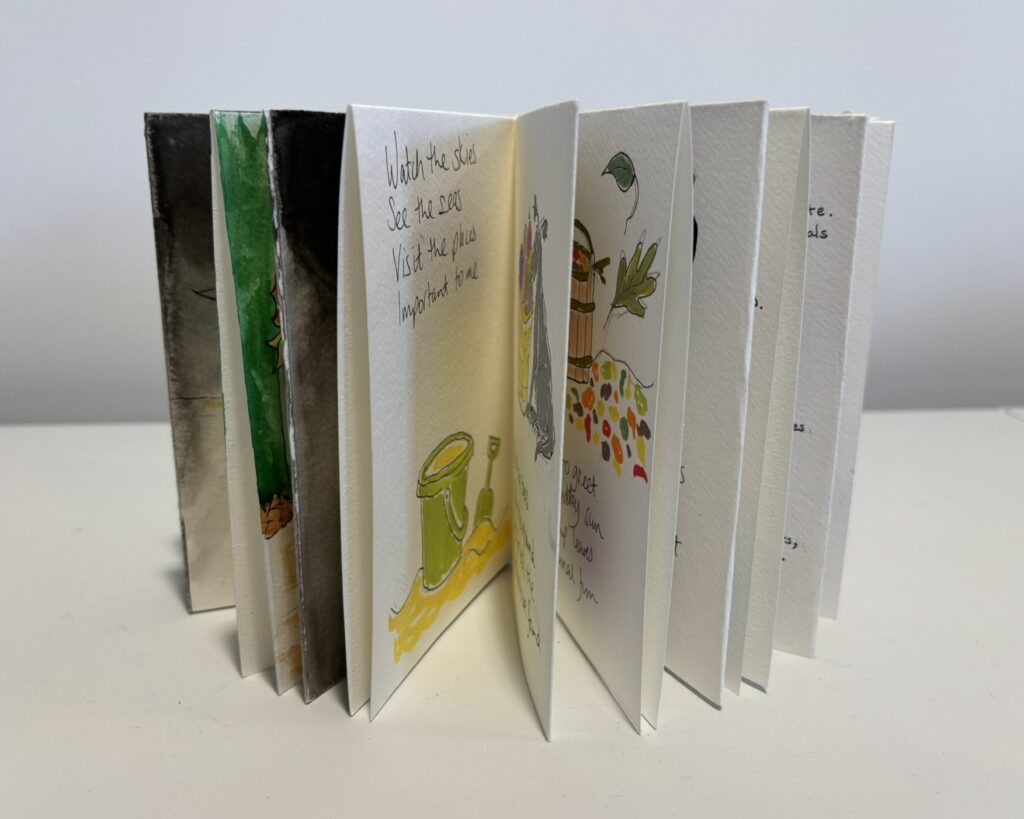 The book is shown standing upright with pages fanned open in an accordion-like arrangement, revealing multiple illustrated spreads. Visible pages include handwritten text, small painted drawings of seaside objects such as a bucket, spade, shells, and leaves, and soft watercolour-style imagery in greens, yellows, and greys. The layered pages create a sculptural silhouette and suggest a narrative or visual journey through memories of the sea and beach.