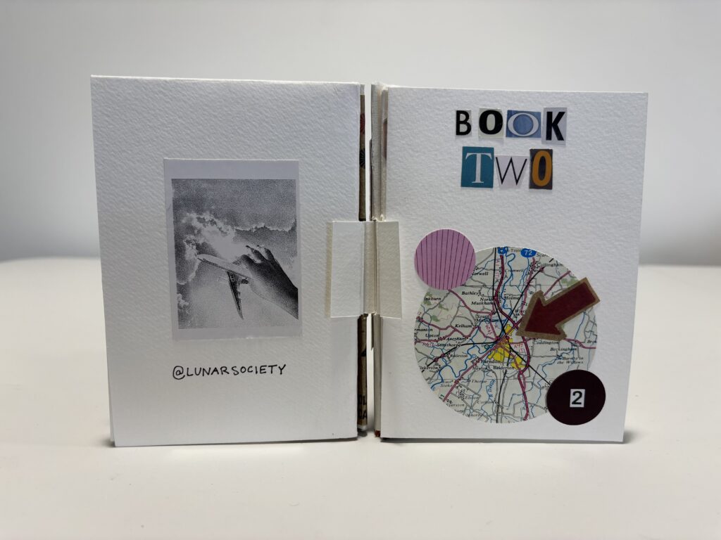 Small handmade artist’s books bound together and displayed upright side by side on a white surface against a plain grey background. The left book has a white textured cover with a small black-and-white photographic image at the centre and the handwritten handle “@LUNARSOCIETY” below. The right book has a white cover titled “BOOK TWO” in cut-out ransom-style letters, decorated with a circular map collage, a pink striped circle, a red arrow shape, and a black circle labelled “2”. Both books have a tactile, crafted appearance with visible folded bindings.