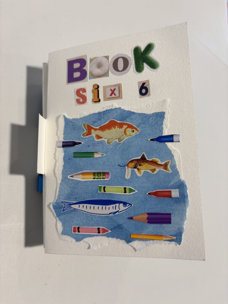 Handmade artist’s book cover titled “BOOK six,” created from collage letters in different fonts and colours. Below the title is a torn-edged blue paper panel resembling water. Across it are cut-out images of fish in various styles – including orange-gold fish and a simple blue line-drawn fish – interspersed with brightly coloured crayons and pencils in purple, green, yellow, red, pink, and orange. The arrangement gives the impression of fish and drawing tools swimming together in water.